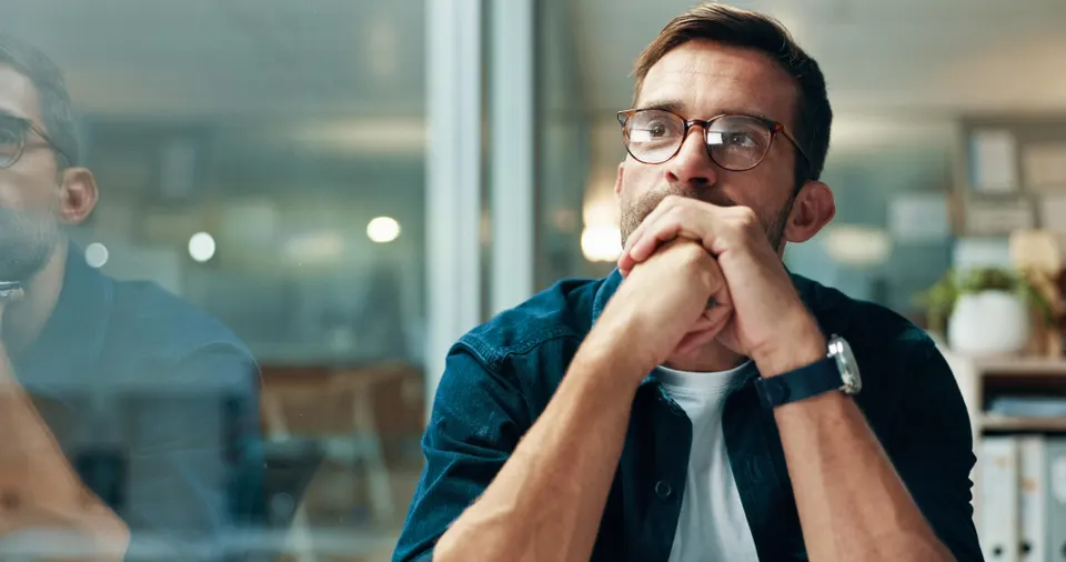 Man with glasses sits at a glass-walled office, hands clasped under chin, looking thoughtful.